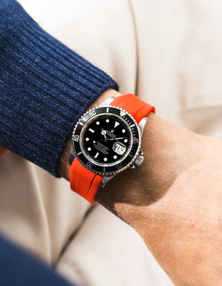 Red FKM rubber strap integrated for original clasp on Rolex Submariner 16610LN full steel, black ceramic bezel, black dial, angled top-down wrist shot, smooth red-orange strap texture, navy blue knit sweater sleeve, cream background