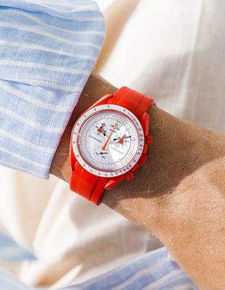 Smooth red rubber strap without texture on Omega x Swatch MoonSwatch Mission to Mars, wrist shot against cream fabric background, white dial with red chronograph hands, matching red bioceramic case and bezel, blue and white striped shirt visible at left