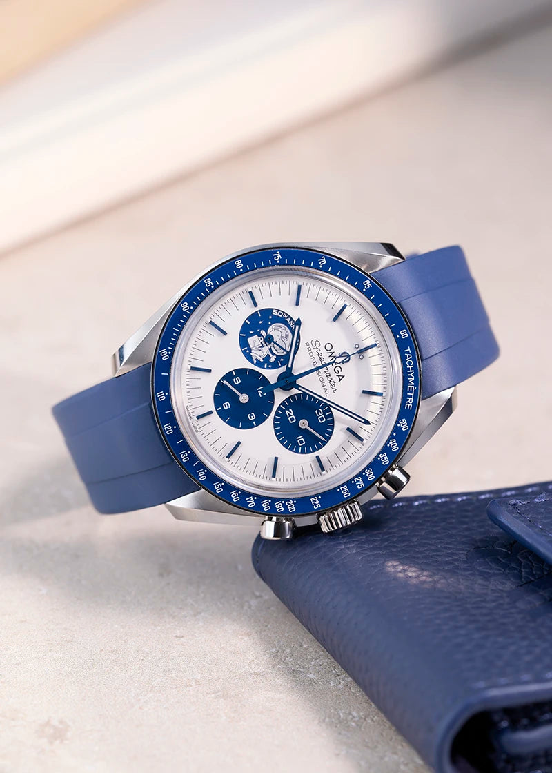 Omega Speedmaster Silver Snoopy Award with blue rubber strap from Wristbuddys, lying flat with the crown resting on a blue leather pouch.