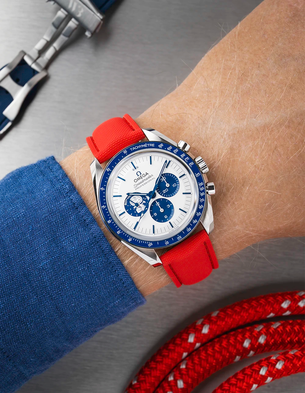 Omega Snoopy Speedmaster with a blue face and red strap on a wrist, with additional straps in the background.