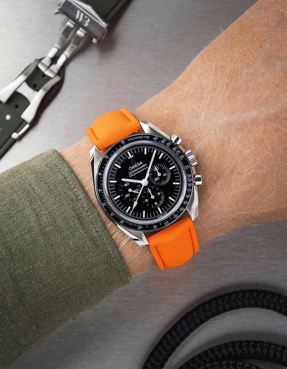 Wrist wearing a black Omega Speedmaster with a orange strap  with a detailed face on a wrist with a green sleeve.