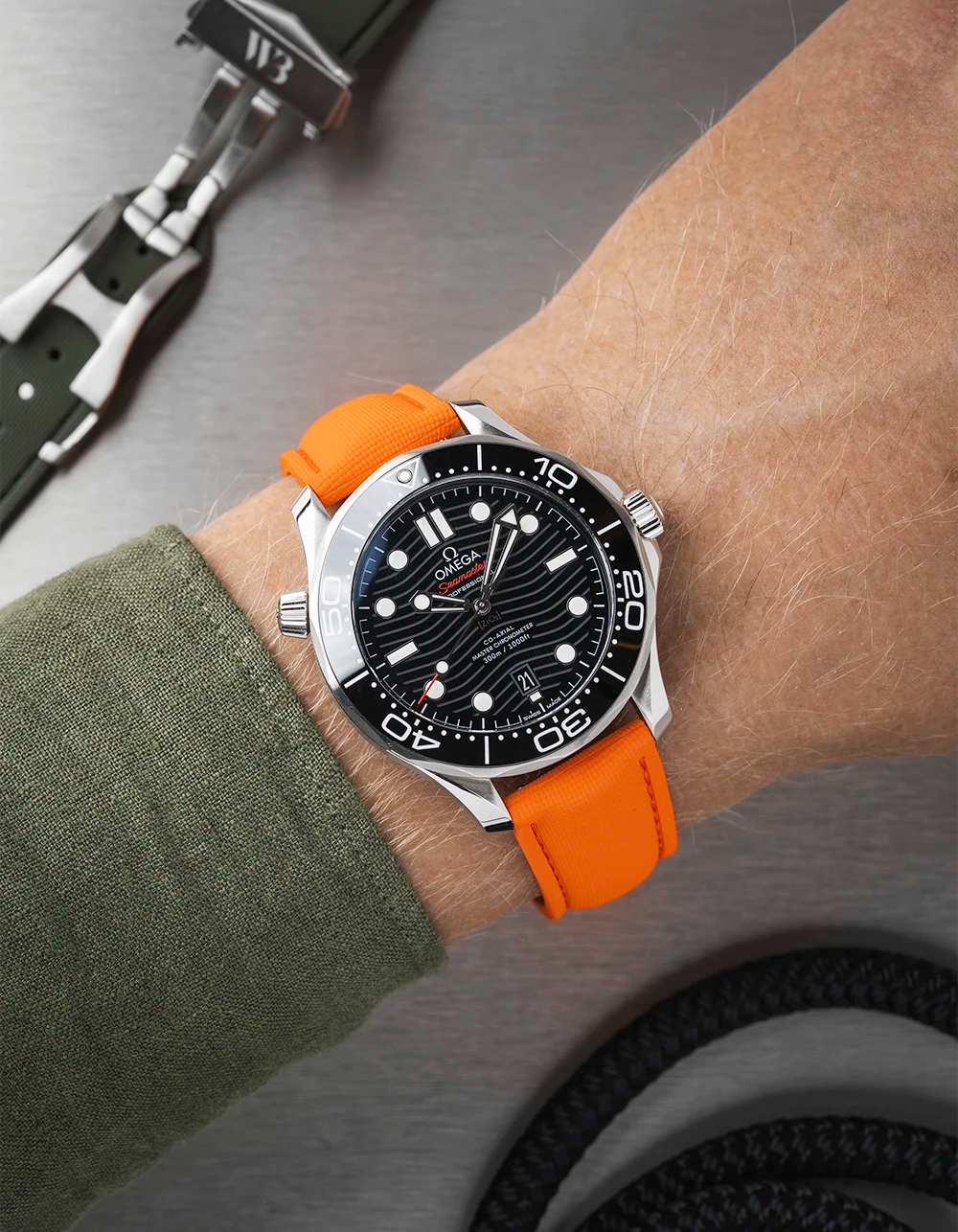Wrist wearing a black Omega Seamaster with a orange strap  with a detailed face on a wrist with a green sleeve.