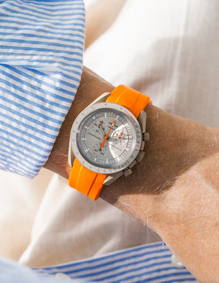 Smooth plain orange rubber strap without texture on Omega x Swatch MoonSwatch Mission to Jupiter, wrist shot against cream fabric background, silver-grey dial with orange chronograph hands, blue and white striped shirt visible at left and lower right