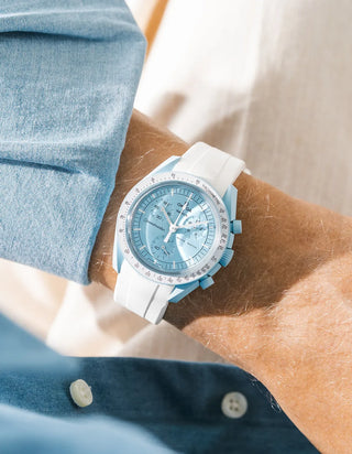 Smooth plain white rubber strap without texture on Omega x Swatch MoonSwatch Mission to Uranus, wrist shot from above against cream background, pale sky blue dial and light blue bioceramic case, white tachymeter bezel, blue chambray shirt with two white buttons visible at lower left