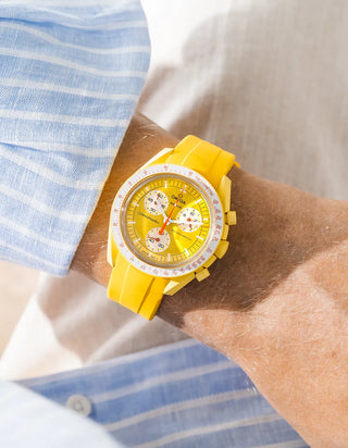 Smooth plain yellow rubber strap without texture on Omega x Swatch MoonSwatch Mission to the Sun, wrist shot from above against cream fabric background, bright yellow dial with red chronograph hands, matching yellow case and white tachymeter bezel, blue and white striped shirt visible at left and lower right with white button