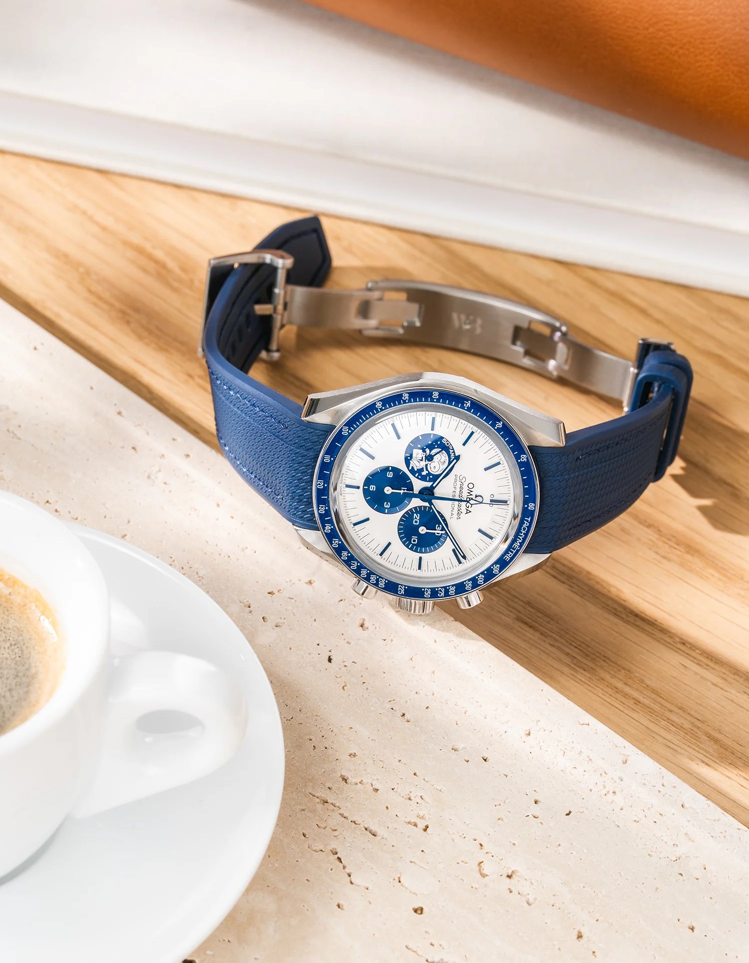 Navy blue Vertech rubber strap for Omega Speedmaster Silver Snoopy Award, fitted on the watch with deployant clasp, displayed on a wooden surface next to an espresso cup.