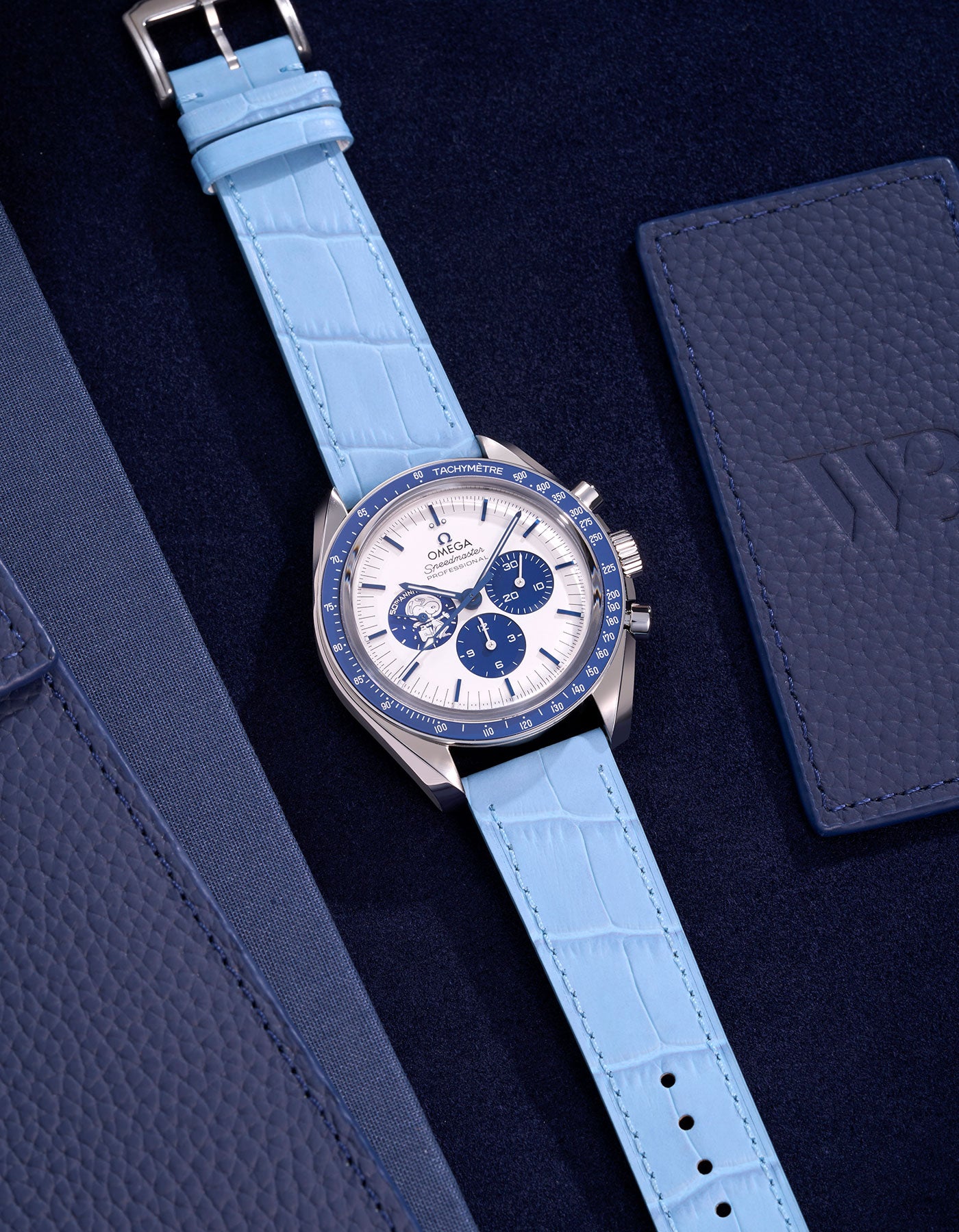 Omega Speedmaster Silver Snoopy Edition watch paired with a light blue alligator leather strap on a dark blue surface