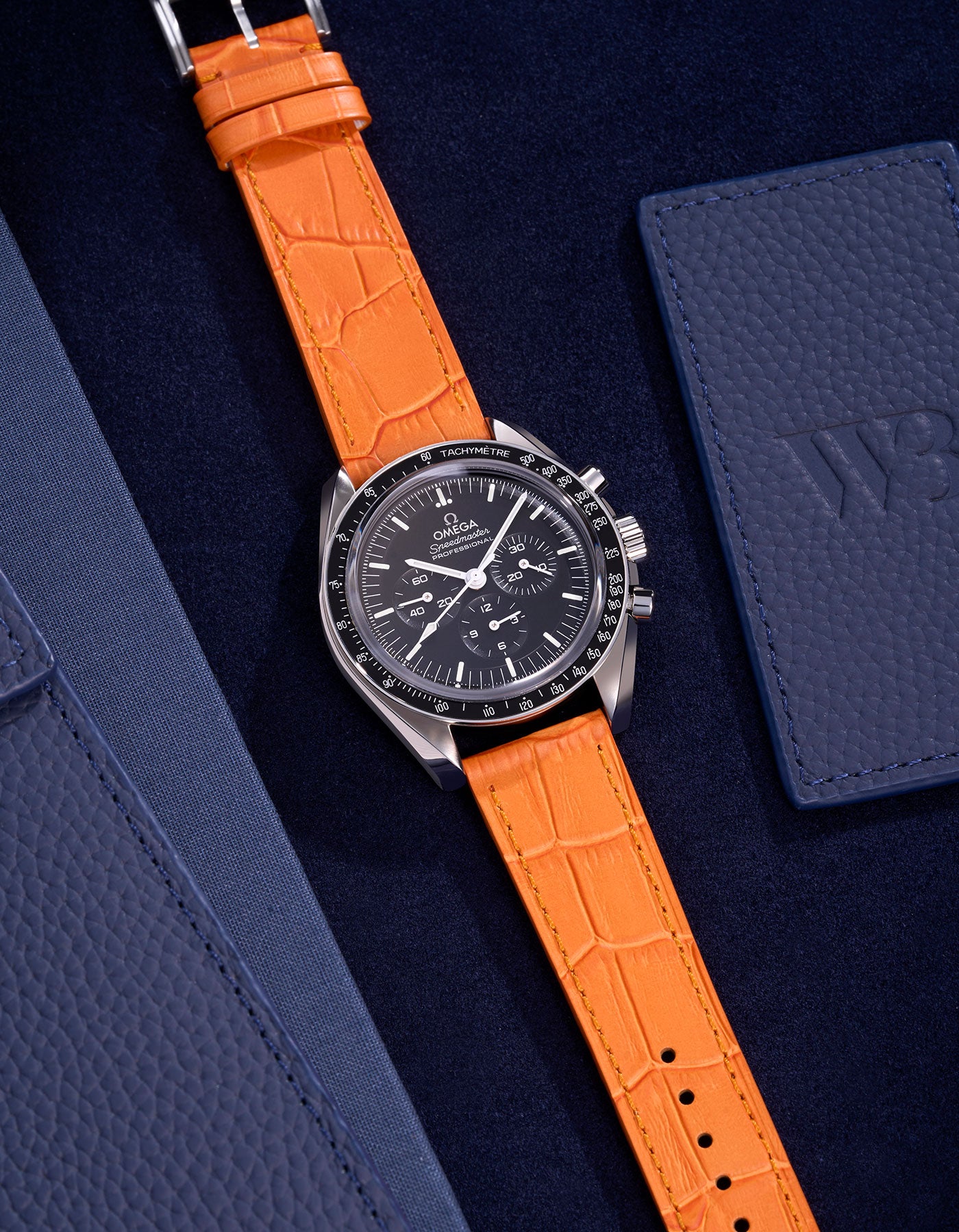 Omega Speedmaster watch with bright orange alligator leather strap on a dark blue background