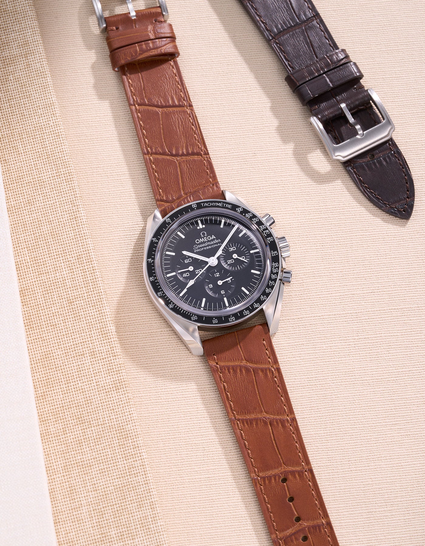 Omega Speedmaster watch with a tan alligator leather strap on a beige background