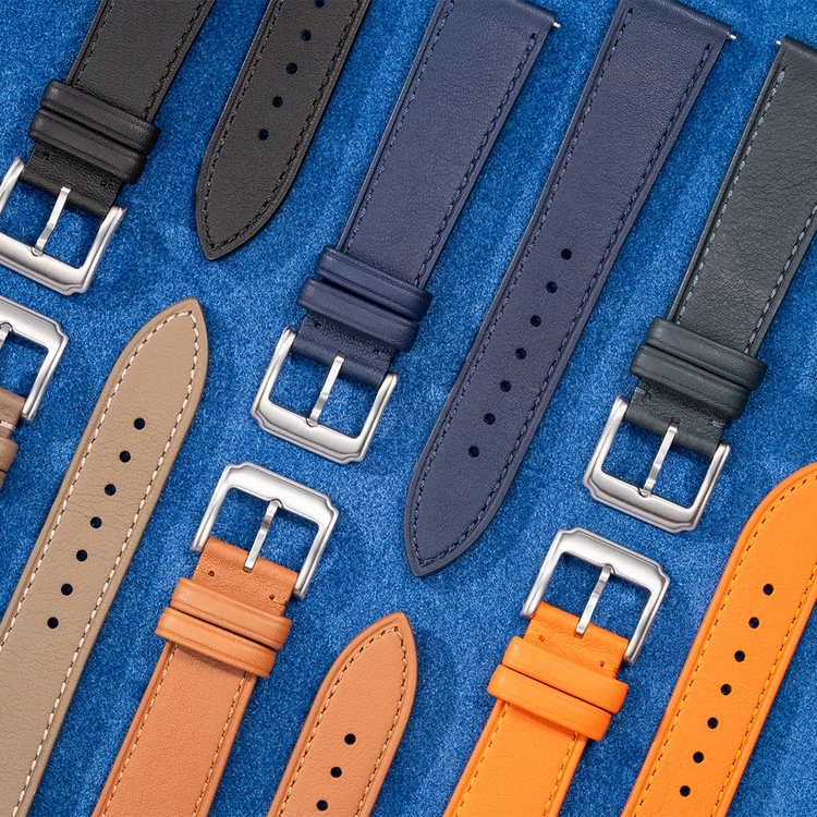 Flat lay of cowhide leather watch straps in multiple colors including black, navy blue, tan, beige, and orange with stainless steel buckles on blue suede background