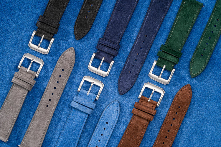Flat lay of suede leather watch straps in multiple colors including black, grey, blue, brown, and green with stainless steel buckles on blue suede background