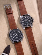 Side-by-side view of Omega Speedmaster and Rolex Submariner watches, both featuring tan alligator leather straps on a beige surface