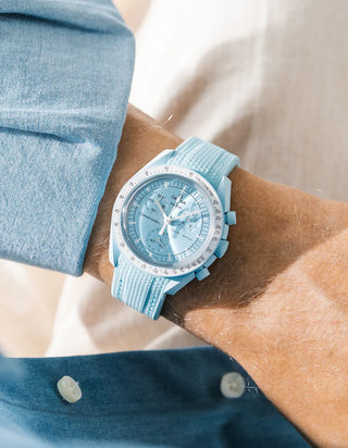 Turquoise rubber strap with vertical ribbed texture on Omega x Swatch MoonSwatch Mission to Uranus, wrist shot against cream fabric background, pale sky blue dial and matching light blue bioceramic case, white tachymeter bezel, blue chambray shirt with two white buttons visible at lower left