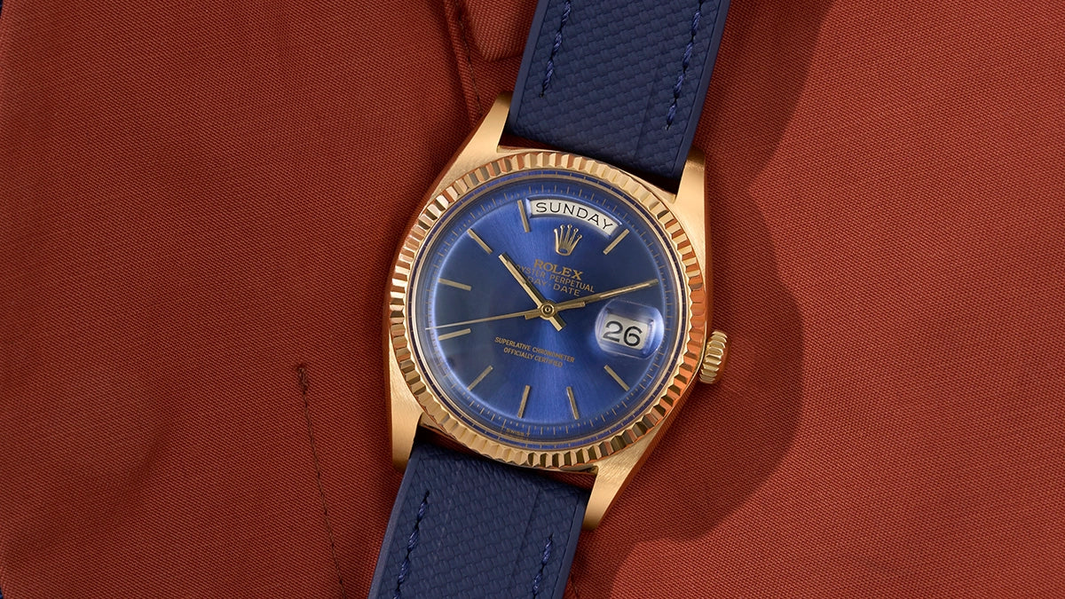 Gold Rolex watch with a blue dial and blue universal strap on a red fabric background.