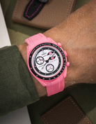 Pink watch with a detailed face on a wrist, worn by a person in a green sleeve.