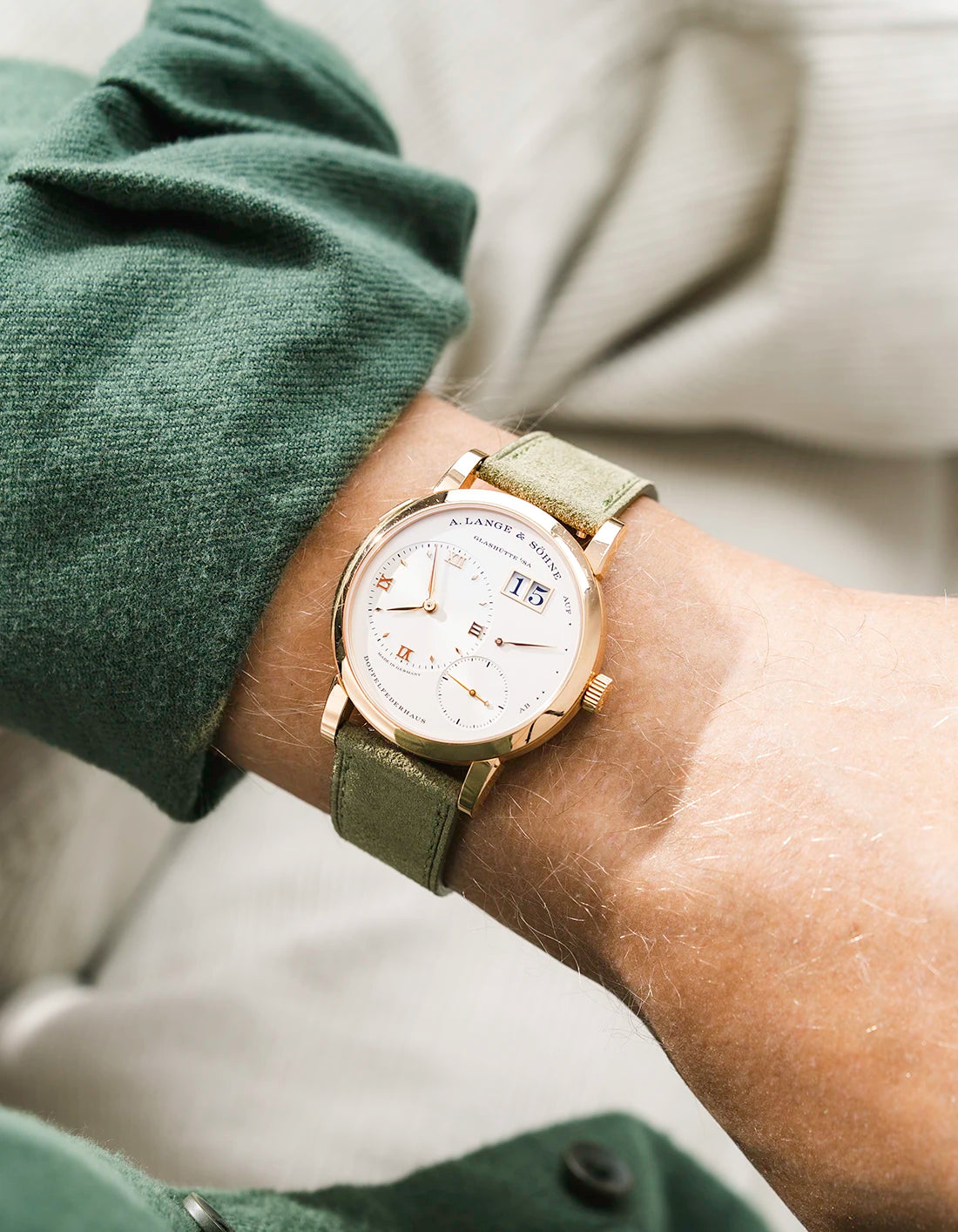 A Lange Shöne in white gold with a herb green suede leather strap from Wristbuddys on a wrist, wearing a green sleeve.