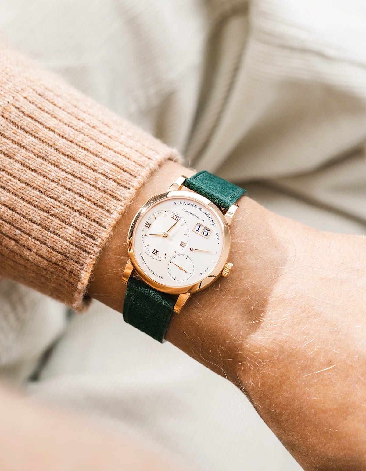 A Lange Shöne in rose gold with a green suede leather strap from Wristbuddys on a wrist, wearing a beige sleeve.