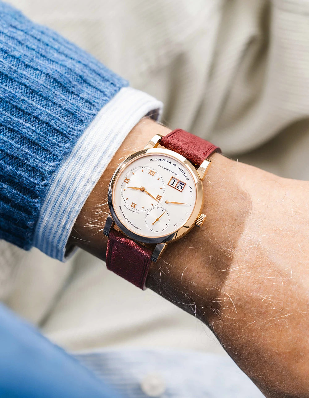 A Lange Shöne in white gold with a Burgundy suede leather strap from Wristbuddys on a wrist, wearing a blue sleeve.