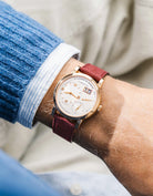 A Lange Shöne in white gold with a Burgundy suede leather strap from Wristbuddys on a wrist, wearing a blue sleeve.