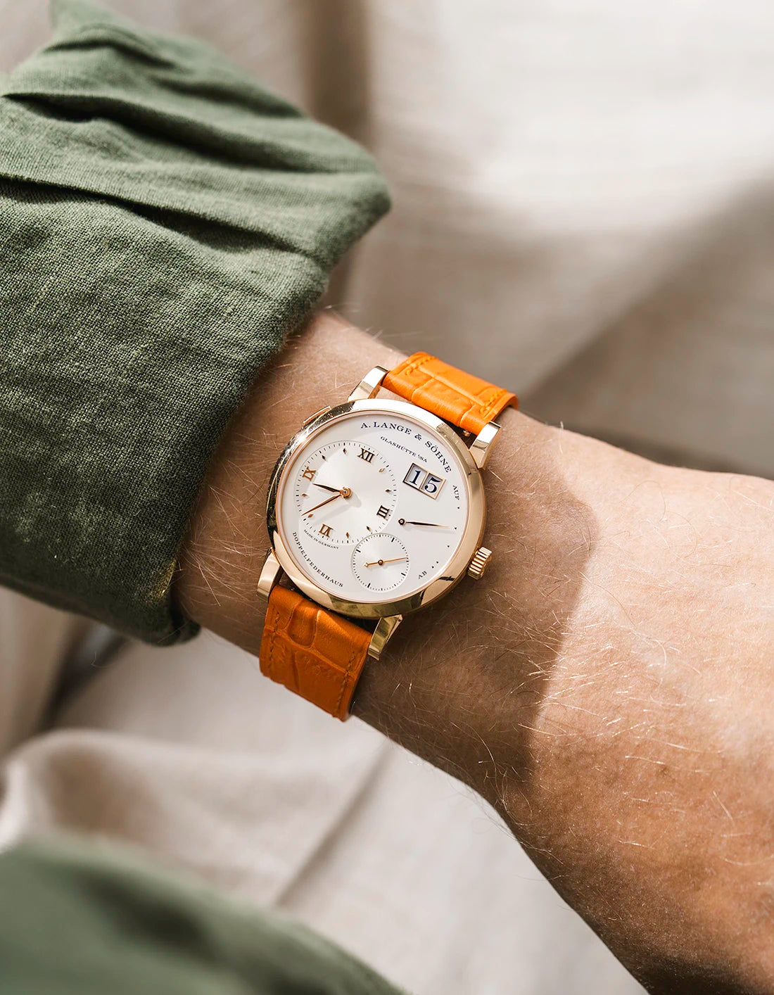 A Lange Shöne in rose gold with a orange embossed alligator leather strap from Wristbuddys on a wrist, wearing a green sleeve.