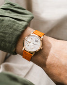 A Lange Shöne in rose gold with a orange embossed alligator leather strap from Wristbuddys on a wrist, wearing a green sleeve.