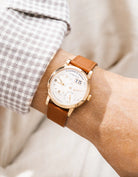 A Lange Shöne in rose gold with a brown calfskin leather watch strap from Wristbuddys on a wrist, wearing a beige sleeve.