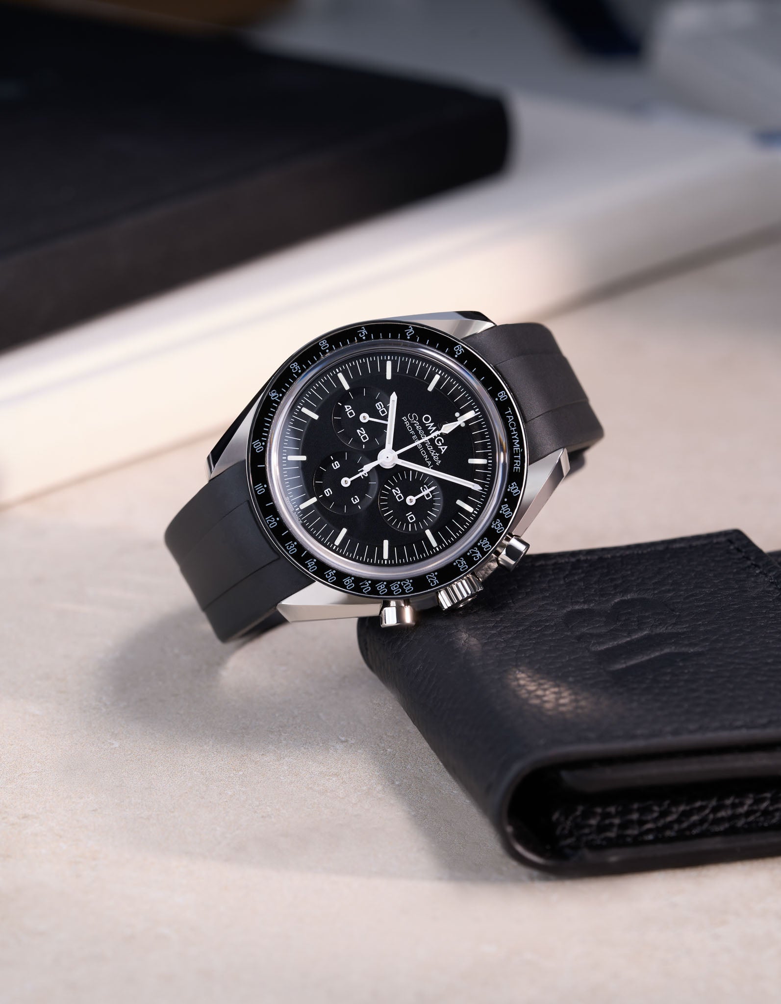 Omega Speedmaster fitted with black FKM rubber strap – angled product photo on beige surface with black leather watch pouch by Wristbuddys