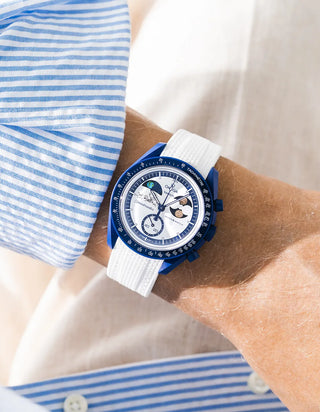 White rubber strap with cross-hatched ribbed texture on Omega x Swatch MoonSwatch Mission to Earthphase Moonshine Gold, wrist shot against cream fabric background, white dial with blue moonphase complication, blue and white striped shirt visible at left and lower left