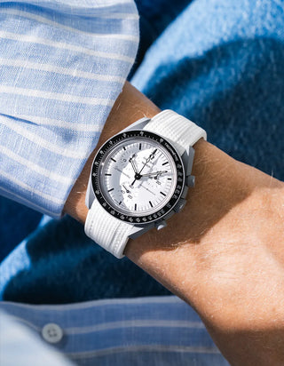 Plain white rubber strap without stitching and fine ribbed texture on Omega x Swatch MoonSwatch Mission to the Moon 1965, wrist shot against blue fluffy textile and blue and white striped shirt background, white dial with black subdials and black tachymeter bezel, white bioceramic case, all-white monochromatic look, white button on shirt visible at lower left