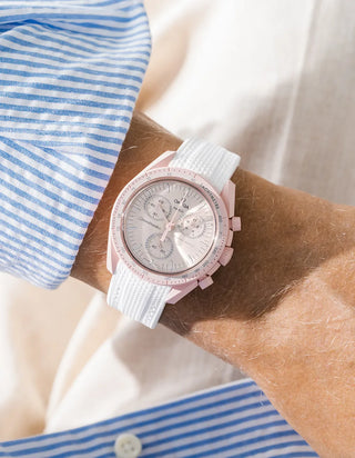 White rubber strap with vertical ribbed texture on Omega x Swatch MoonSwatch Mission to Venus, wrist shot against cream fabric background, pale pink dial and pink bioceramic case and bezel, blue and white striped shirt with white button visible on both sides