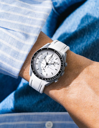 White rubber strap with black contrast stitching and fine ribbed texture on Omega x Swatch MoonSwatch Mission to the Moon 1965, wrist shot against blue fluffy textile and blue and white striped shirt background, white dial with black subdials and black tachymeter bezel, white bioceramic case, all-white monochromatic look with black stitching as sole contrast detail