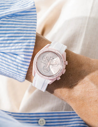 White rubber strap with pink contrast stitching and vertical ribbed texture on Omega x Swatch MoonSwatch Mission to Venus, wrist shot against cream fabric background, pale pink dial and pink bioceramic case and bezel, blue and white striped shirt with white button visible at left and lower right