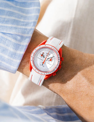 White rubber strap with red contrast stitching on Omega x Swatch MoonSwatch Mission to Mars, wrist shot against cream fabric background, white dial with red-orange chronograph hands, bright red-orange bioceramic case and bezel, blue and white striped shirt visible at left