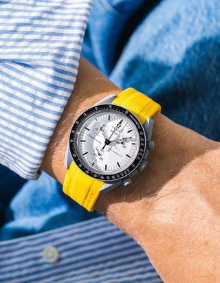 Bright yellow rubber strap with fine diagonal ribbed texture on Omega x Swatch MoonSwatch Mission to the Moon 1965, wrist shot against blue denim jacket background, white dial with black subdials and black tachymeter bezel, white bioceramic case, blue and white striped shirt with white button visible at lower left