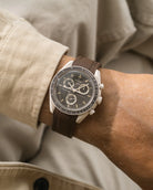 Close-up of a wrist wearing a watch with a brown rubber strap, against a neutral background.