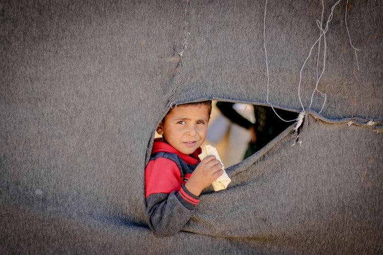 A child peeking through a torn shelter wall while eating bread, representing the strength and hope that Feed a Buddy brings to communities facing hunger.