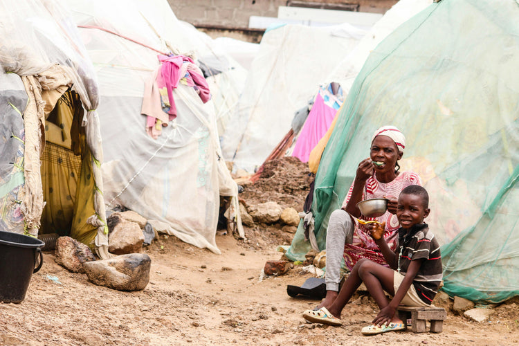 A woman and a child eating outside makeshift shelters, representing the mission of Feed a Buddy to fight hunger and provide support.