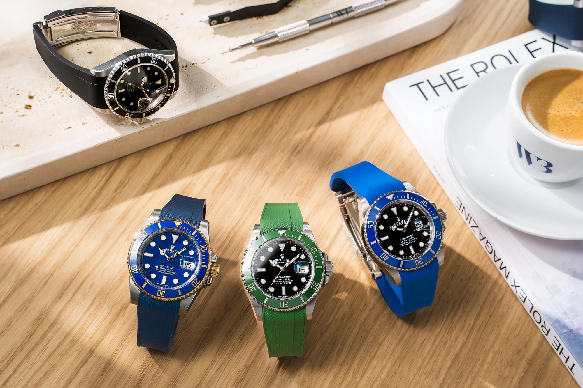 Multiple Rolex Submariner rubber straps in black, blue and green on watches displayed with Rolex Magazine and coffee