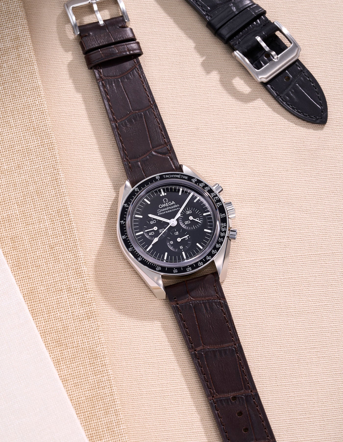 Omega Speedmaster watch with a rich brown alligator leather strap on a beige background