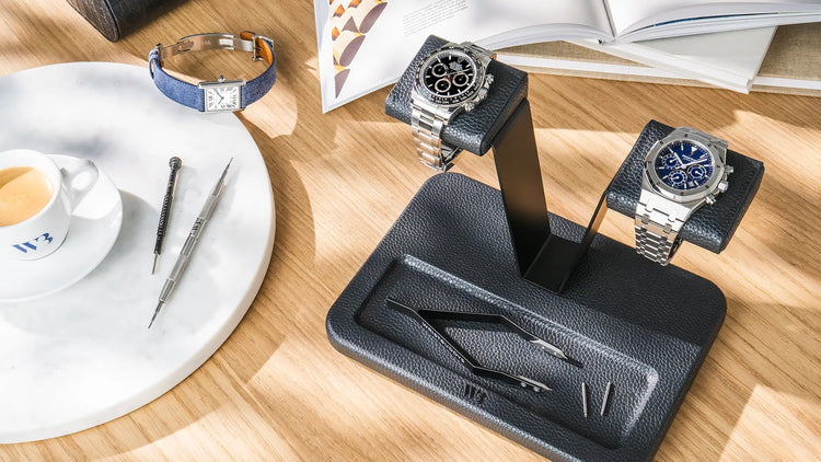 A black leather double watch stand displaying luxury timepieces on a wooden desk